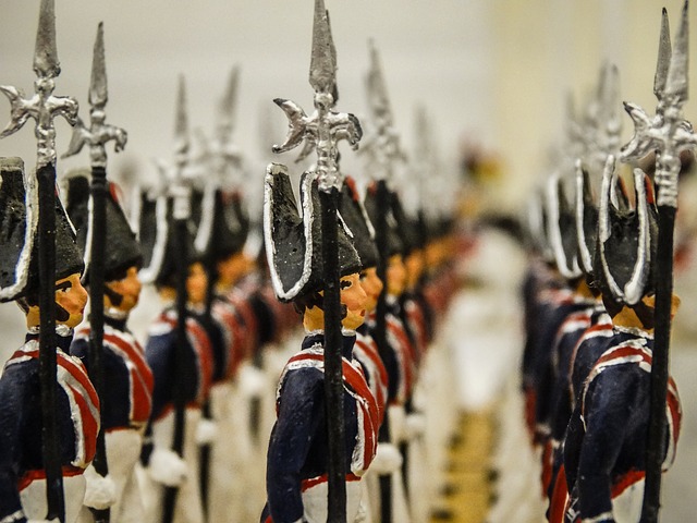 Zbliżenie na szeregi pomalowanych figurek ołowianych przedstawiających żołnierzy piechoty z przełomu XVIII i XIX wieku. Żołnierze w ciemnoniebieskich mundurach i czarnych czapkach (bicornach) trzymają pionowo długie halabardy lub lance ze srebrnymi grotami.