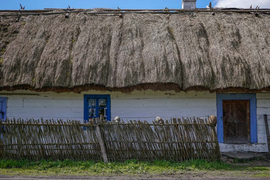 Zbliżenie na białą, drewnianą chatę z dachem krytym grubą strzechą. Dom ma niebieskie obramowania okien i drzwi, a przed nim stoi płot z plecionych witek.