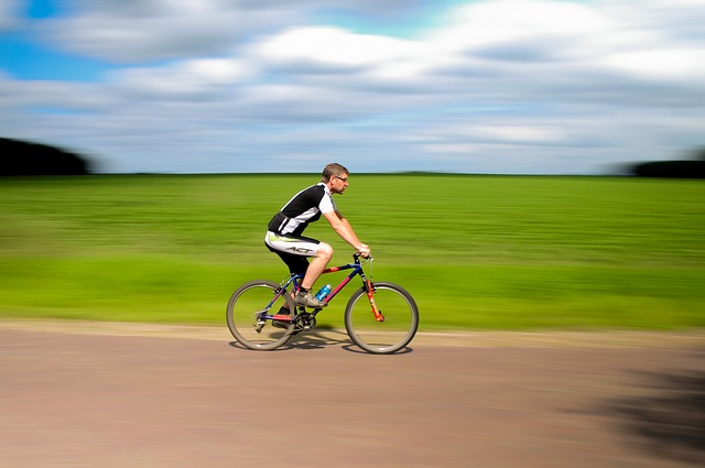 Dynamiczne ujęcie w technice panoramowania, przedstawiające rowerzystę jadącego na rowerze górskim po wiejskiej drodze w jasny, częściowo zachmurzony dzień.