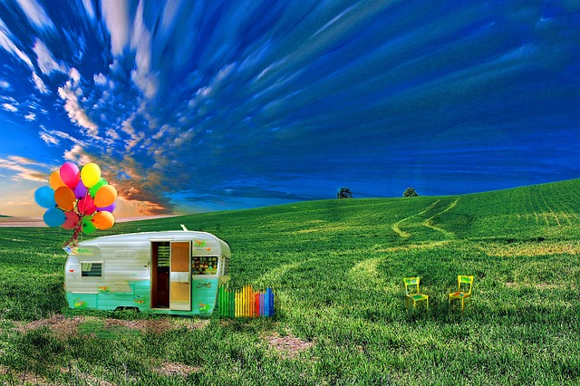 Surrealistyczna grafika przedstawiająca zabytkową przyczepę kempingową na rozległym, intensywnie zielonym polu. Do przyczepy przywiązany jest duży pęk kolorowych balonów z helem. Po prawej stronie stoją dwa puste, jaskrawożółte krzesła. Niebo nad scenerią jest nienaturalnie niebieskie i dynamiczne, przypominające ruch.