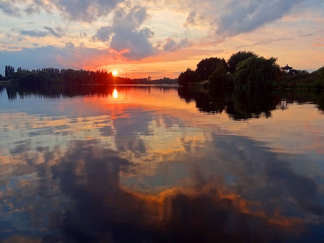 Zachód słońca nad spokojnym jeziorem, z gładką taflą wody na pierwszym planie, w której odbijają się barwy nieba i sylwetki drzew na brzegach po lewej i prawej stronie.