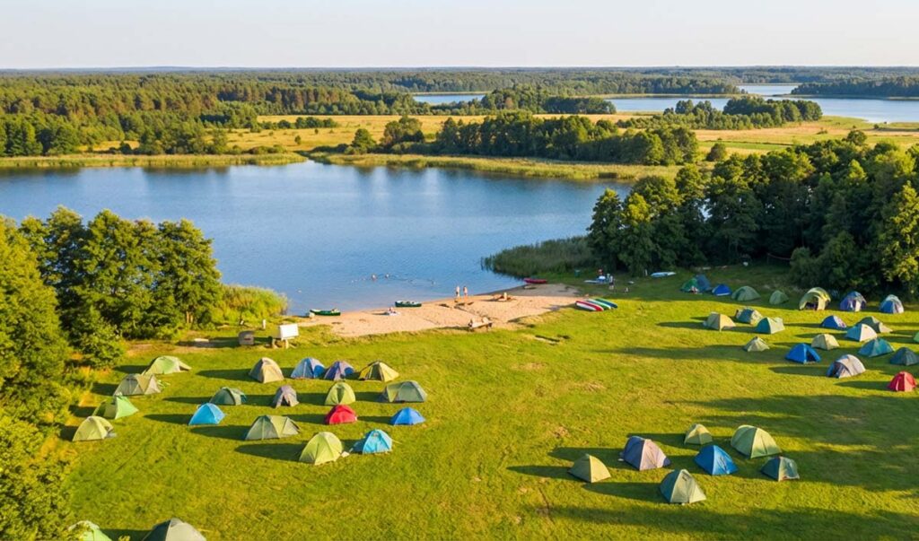 Szeroka panorama z lotu ptaka na zielone pole namiotowe z dziesiątkami kolorowych namiotów. Pole sąsiaduje z piaszczystą plażą i czystym jeziorem w otoczeniu lasów.