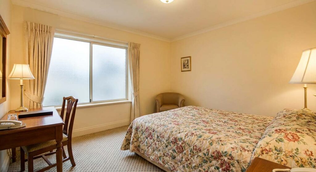 Przytulny pokój hotelowy o klasycznym wystroju. W pokoju znajduje się łóżko przykryte kwiecistą narzutą, drewniane biurko z krzesłem przy oknie oraz fotel w rogu. Wnętrze jest jasne i oświetlone ciepłym światłem lamp stołowych.