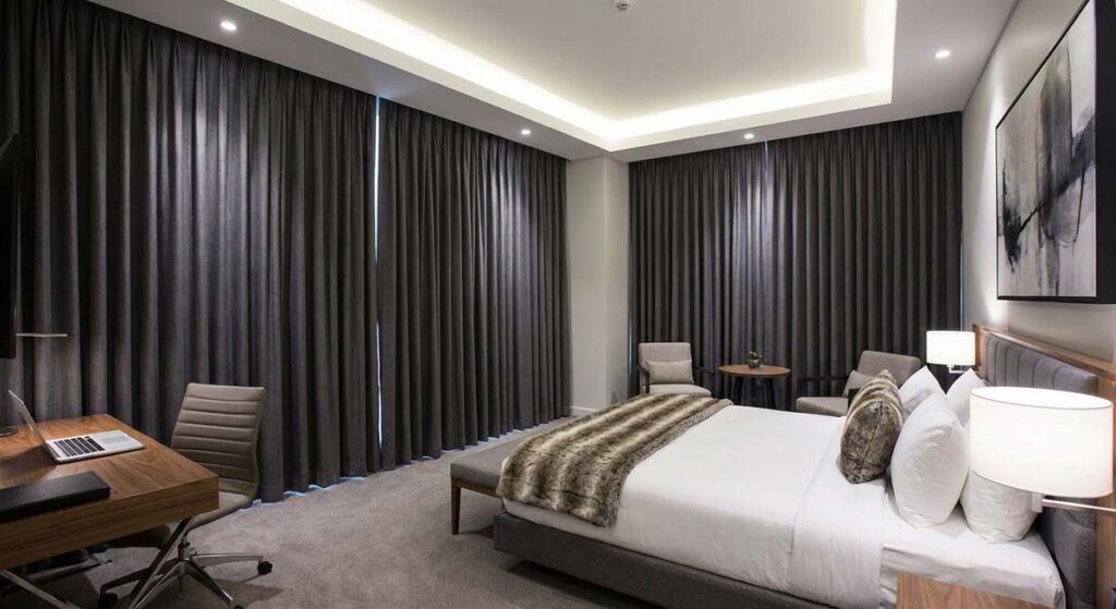 Nowoczesny i elegancki apartament hotelowy w ciemniejszej tonacji. Duże łóżko z białym zagłówkiem i futrzanym narzutem, stylowe szare zasłony od sufitu do podłogi oraz minimalistyczne biurko z laptopem. Oświetlenie sufitowe typu LED tworzy luksusowy klimat.