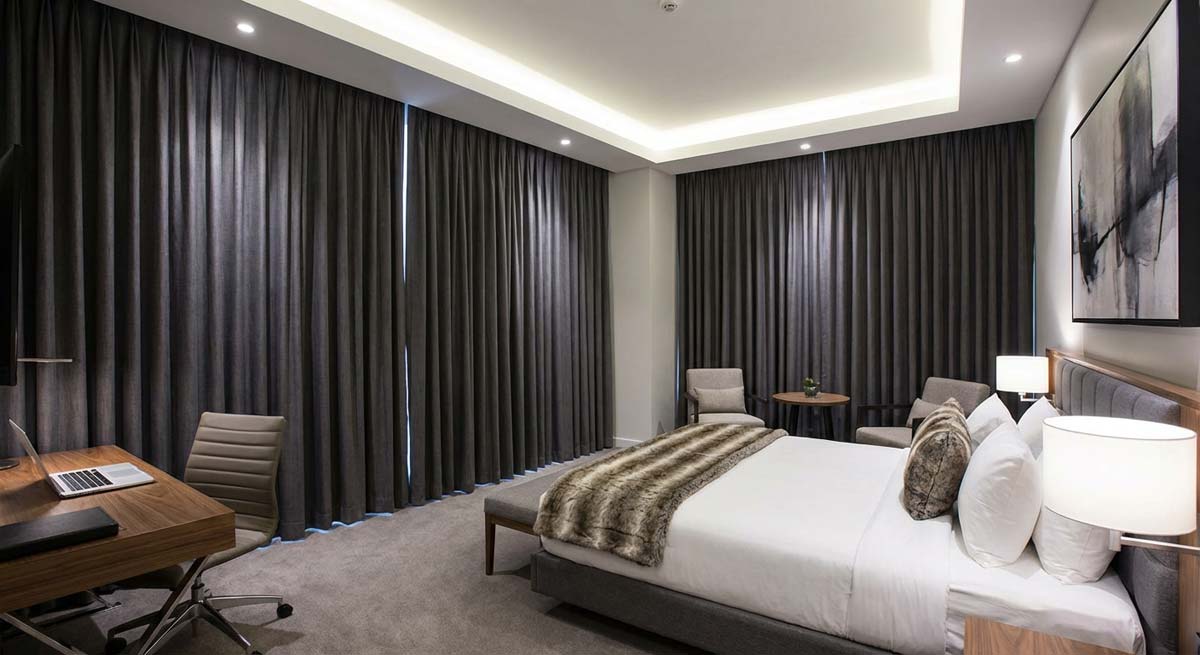 Nowoczesny i elegancki apartament hotelowy w ciemniejszej tonacji. Duże łóżko z białym zagłówkiem i futrzanym narzutem, stylowe szare zasłony od sufitu do podłogi oraz minimalistyczne biurko z laptopem. Oświetlenie sufitowe typu LED tworzy luksusowy klimat.