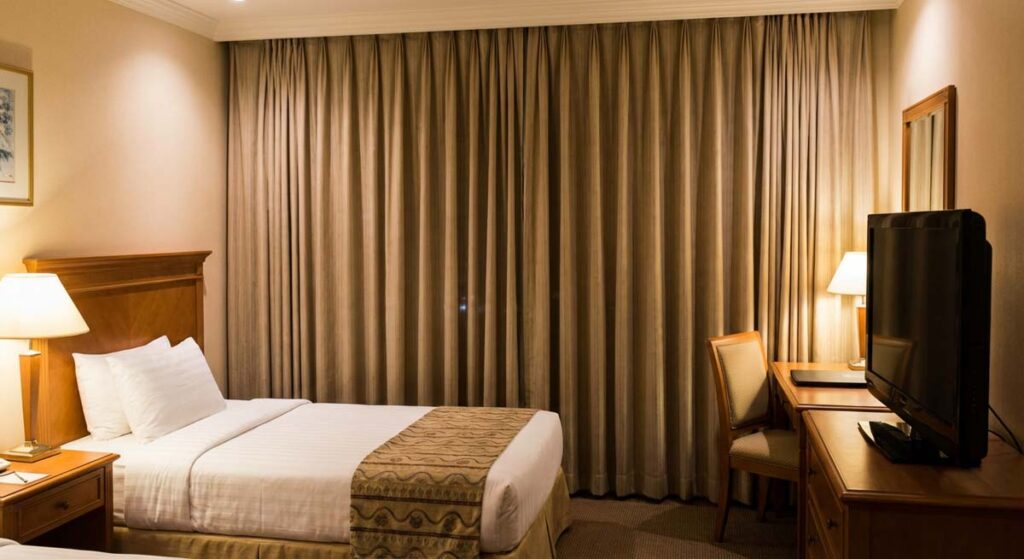 Pokój hotelowy z dwoma łóżkami, utrzymany w ciepłych brązach i beżach. Na pierwszym planie łóżko z białą pościelą, w tle drewniana komoda z telewizorem oraz biurko do pracy.